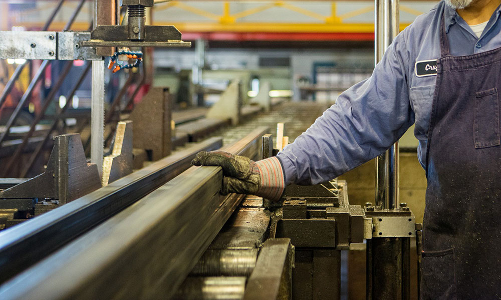 A steel factory worker in a blue jumpsuit and protective glove pushes a steel I-beam along a machine inside a steel factory